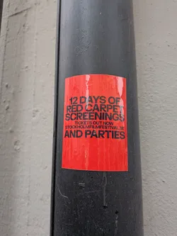 Street sticker A rectangular sticker with a vibrant red background and bold black text. The sticker advertises '12 DAYS OF RED CARPET SCREENINGS' and 'AND PARTIES', with a smaller line stating 'TICKETS OUT NOW' and a website 'STOCKHOLMFILMFESTIVAL.SE'. The sticker appears slightly worn with some minor creases and water marks, adhered to a dark grey or black pipe.