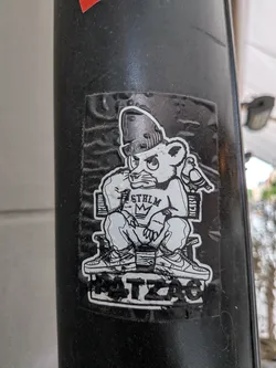 Street sticker A monochromatic sticker featuring a stylized, somewhat grumpy-looking bear-like character. The character wears a beanie, a t-shirt with 'STHLM' and a crown (MK) graphic, baggy pants, and detailed sneakers (resembling Nike Air Force 1s). It is seated on a simple bench, with a small pigeon perched on the back of the bench next to it. Below the bench, the word 'HATZAC' is displayed in a blocky, distressed font. The sticker has a white outline and shows signs of wear, including creases and dirt, indicating it's a physical sticker on a textured black pole.