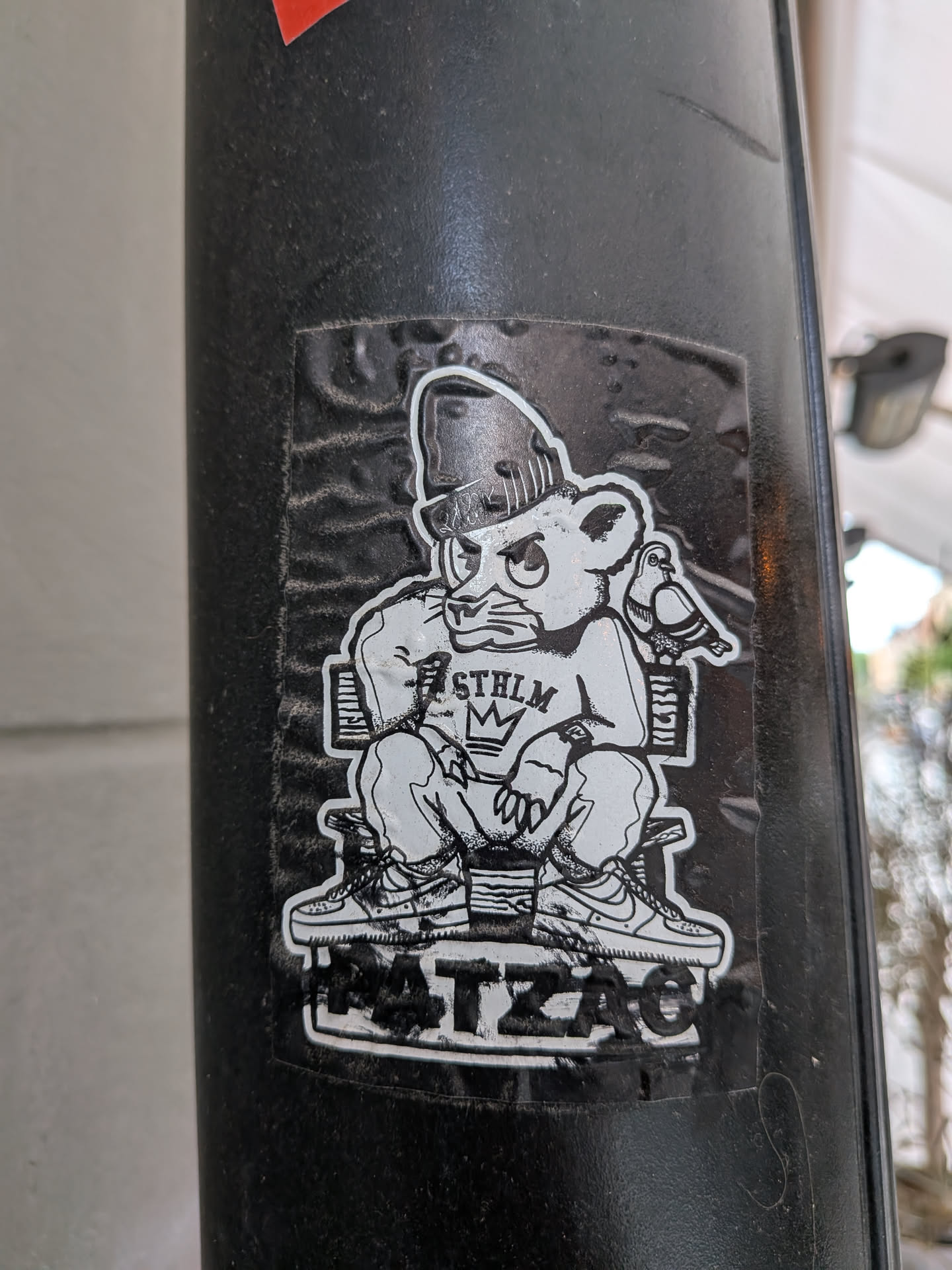 A monochromatic sticker featuring a stylized, somewhat grumpy-looking bear-like character. The character wears a beanie, a t-shirt with 'STHLM' and a crown (MK) graphic, baggy pants, and detailed sneakers (resembling Nike Air Force 1s). It is seated on a simple bench, with a small pigeon perched on the back of the bench next to it. Below the bench, the word 'HATZAC' is displayed in a blocky, distressed font. The sticker has a white outline and shows signs of wear, including creases and dirt, indicating it's a physical sticker on a textured black pole.