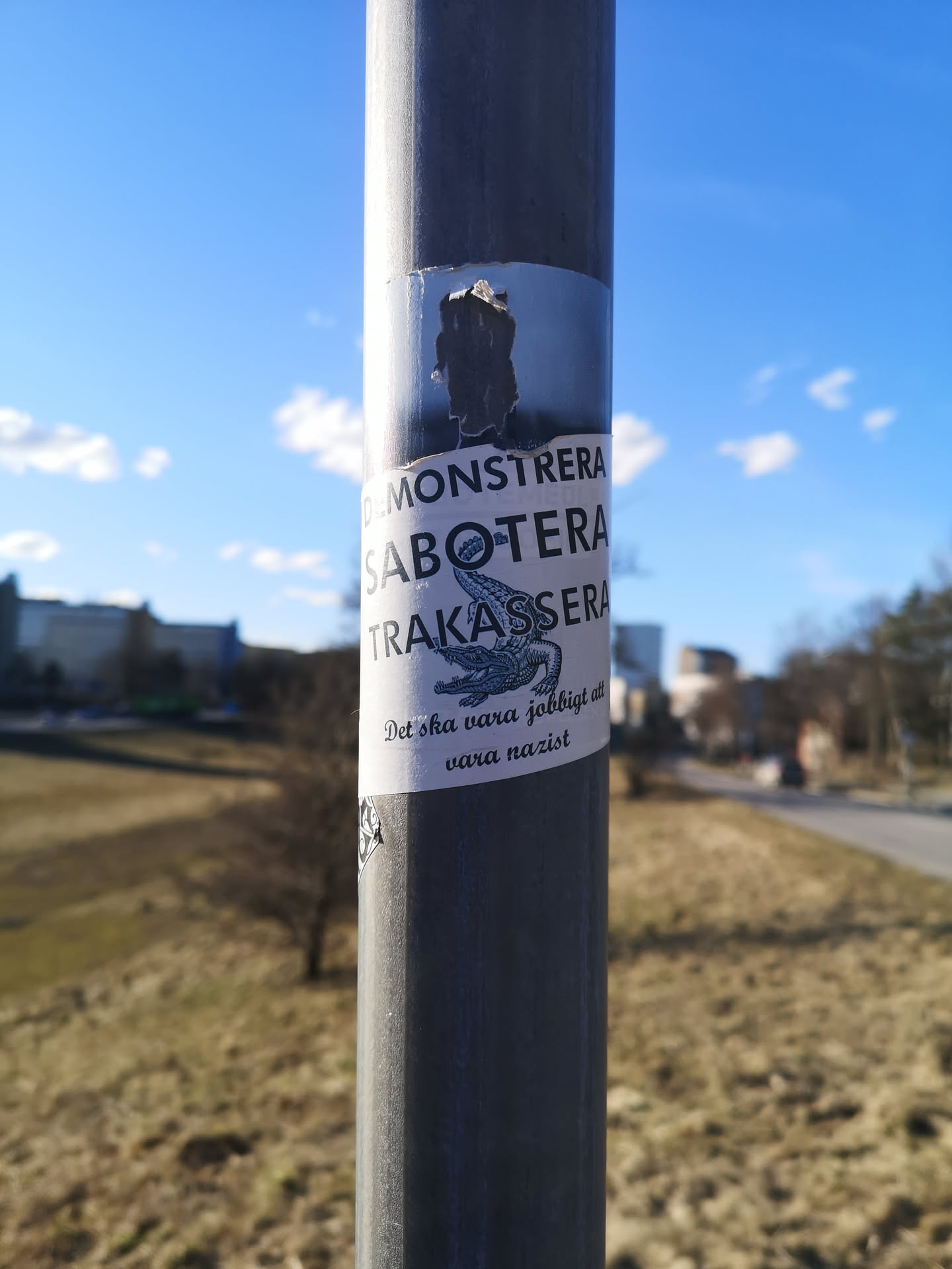 The sticker is rectangular with a white background, adhered to a grey metal pole. It features bold black text in Swedish, arranged vertically, with an illustration of a stylized crocodile or alligator below the main text. The crocodile is depicted in black outline and shading, appearing aggressive with its mouth open. Below the crocodile, there is more text in a smaller, italicized font. The top edge of the sticker is torn and partially obscured by another, older sticker or residue. The overall aesthetic is stark and impactful, using only black and white.