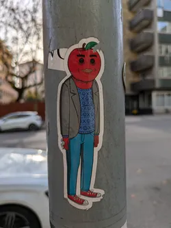 Street sticker A sticker featuring an anthropomorphic apple character wearing a gray jacket, blue jeans, and red sneakers.  The apple head has a smirk and simple eyes. The design is simple and cartoonish.
