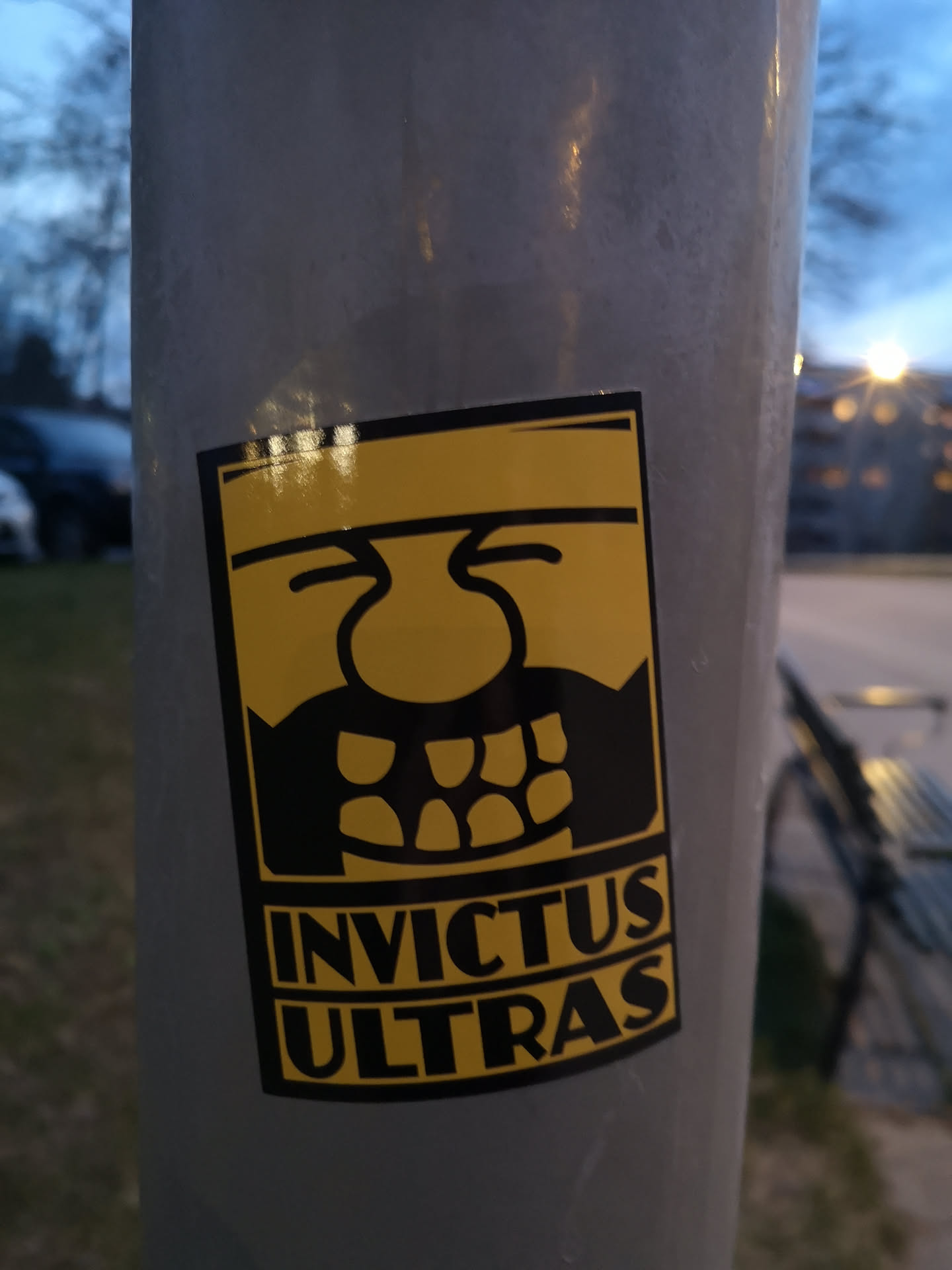 A rectangular sticker featuring a bold, graphic design in black and yellow. The central image is a stylized, somewhat grotesque or monstrous face rendered in black against a bright yellow background. The face has a large, bulbous nose, two simple curved lines for eyes, and a wide, toothy grin revealing multiple irregular teeth. Below the face, the words "INVICTUS ULTRAS" are stacked in two lines, written in bold, black, sans-serif capital letters. The entire design is framed by a thin black border.