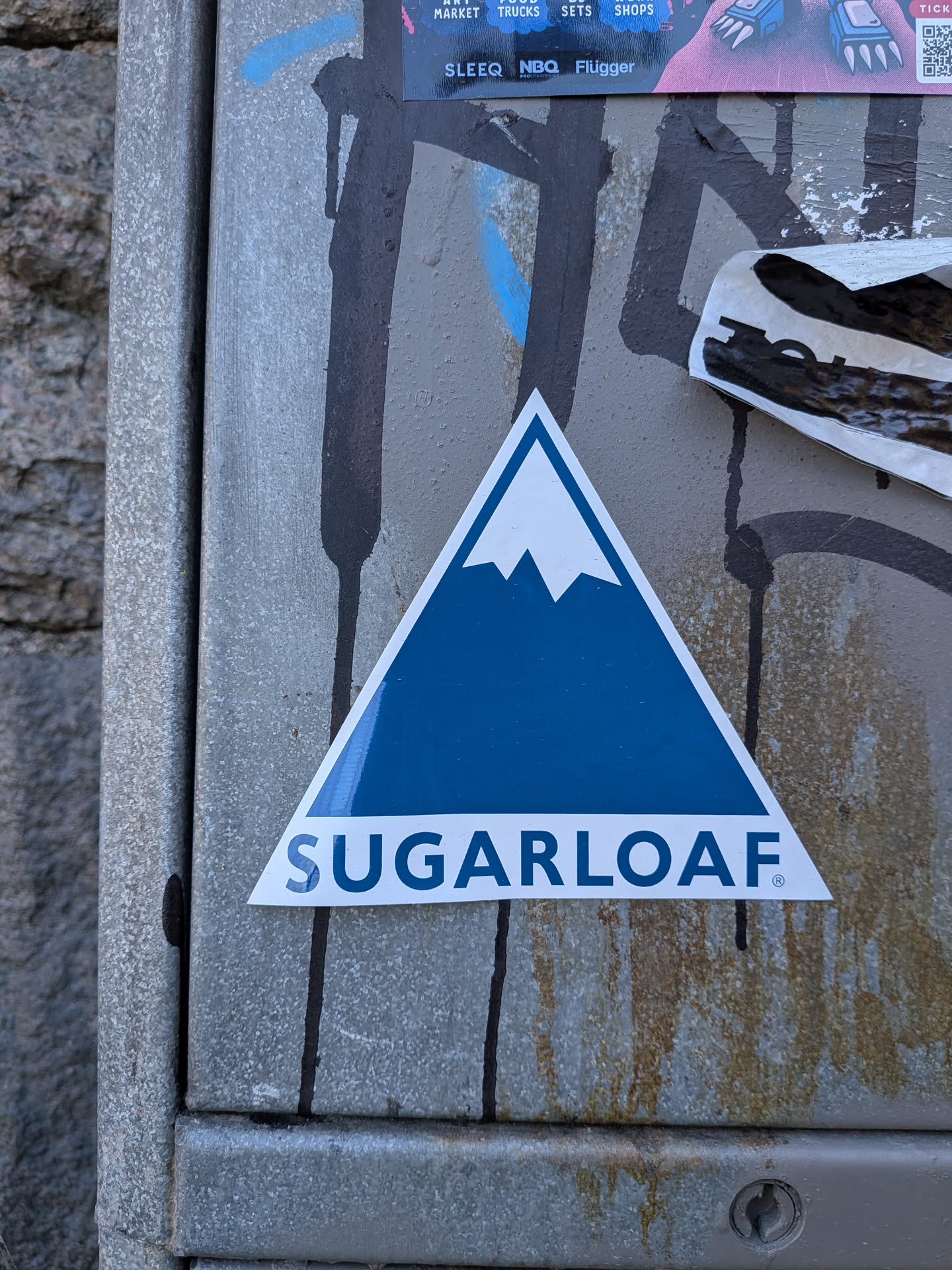 The sticker is a prominent triangular design with a white border. The main body of the triangle is a solid, vibrant blue, representing a mountain. At the apex of this blue triangle, a jagged white shape depicts a snow-capped peak. Below the mountain graphic, the word 'SUGARLOAF' is written in bold, white, sans-serif capital letters, followed by a small registered trademark symbol '®'. The overall appearance is clean, modern, and minimalist, effectively conveying a mountain or ski resort theme.