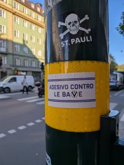 A rectangular white sticker with purple text and double horizontal borders. It features the Italian phrase 'ADESIVO CONTRO LE BAVE' with a stylized 'V' enclosing a star.