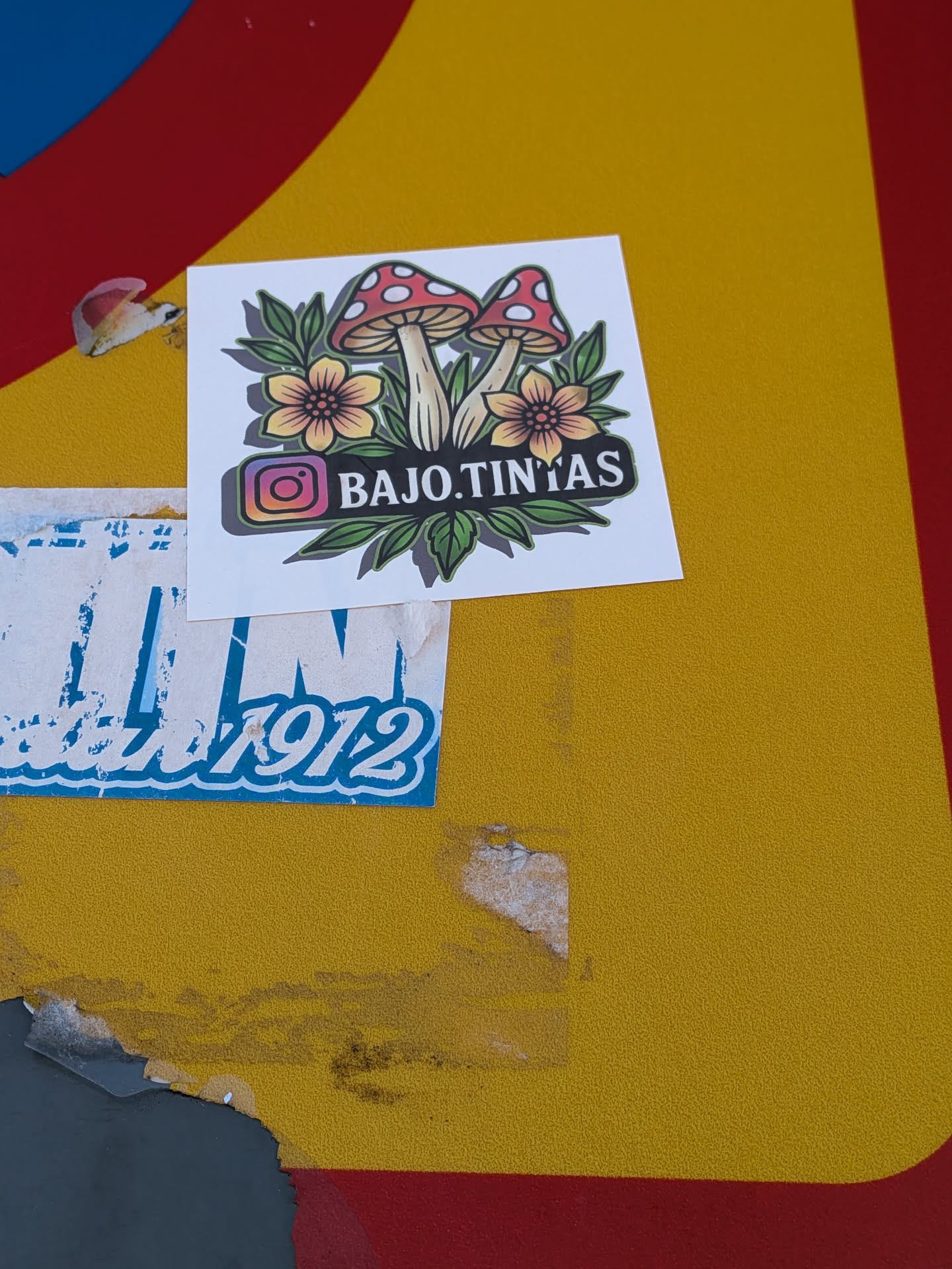 The sticker is square-shaped with a white background. It features a central illustration of two red and white spotted mushrooms with green stems and leaves. Flanking the mushrooms are two stylized yellow flowers with orange centers and green leaves. Below the illustration, a black banner with a purple and green gradient outline contains the text 'BAJO.TINTAS' and an Instagram logo. The overall style is illustrative and somewhat whimsical.