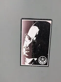 Street sticker This sticker displays a black and white portrait of Malcolm X, with a section of the face rendered in a halftone pattern. A symbol with a star and crescent moon is located in the bottom right corner, possibly representing an organization or movement associated with Malcolm X.
