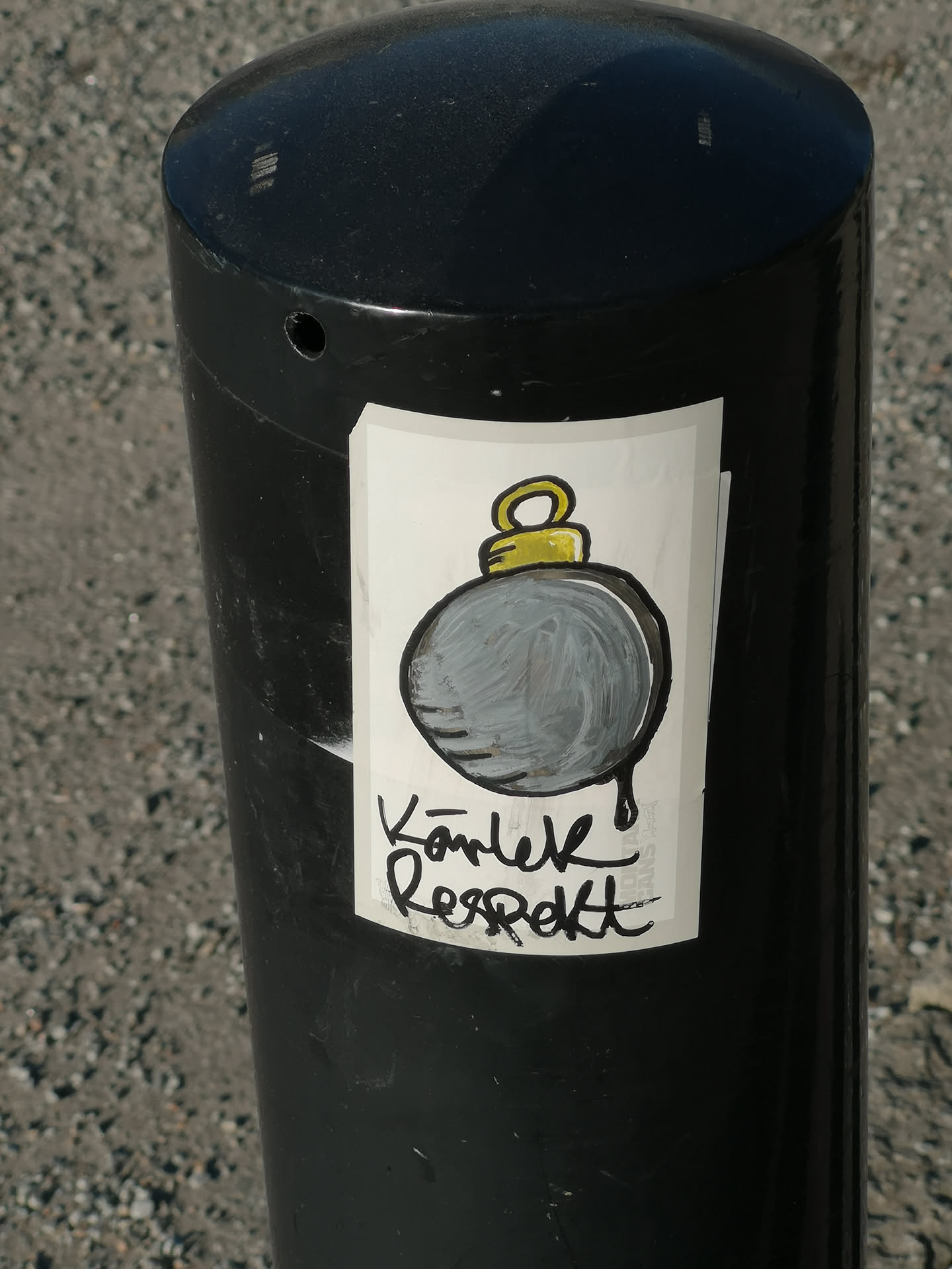 A rectangular sticker with a white background, featuring a stylized, cartoon-like bomb. The bomb is a dark grey sphere with visible brushstrokes, outlined in thick black, and has a bright yellow fuse loop at the top, also outlined in black. A black drip extends from the bottom of the bomb. Below the bomb, in a bold, handwritten-style black script, are the words "Kärlek Respekt". The sticker is placed on a black, cylindrical bollard and shows slight wear and curling at the edges.