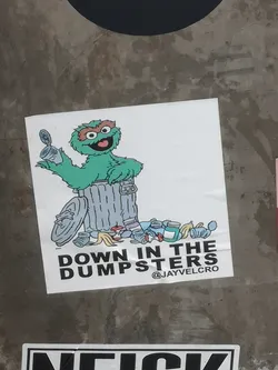 Square white sticker depicting Oscar the Grouch in a trash can holding a tin can, surrounded by litter. Bold black text below reads 'DOWN IN THE DUMPSTERS' with the handle '@JAYVELCRO'.