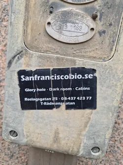 A sticker on a dirty electrical box in Stockholm, Sweden.  The sticker advertises San Fransisco Bio, listing its services (Glory hole, Dark room, Cabins) and contact information.