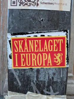 The sticker is rectangular with a bright red background and a yellow border. Prominently displayed in large, bold yellow sans-serif font are the words 'SKÅNELAGET' on the top line and 'I EUROPA' on the bottom line. To the right of the text, there is a yellow emblem of a rampant lion, a common heraldic symbol. The sticker appears to be slightly worn around the edges and is affixed to a dark, textured surface.