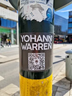 A black and white QR code sticker with the name "YOHANN WARREN" written in white uppercase letters on top of it. The sticker is attached to a yellow and dark green pole on a city street.