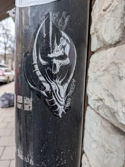 Street sticker The sticker is an irregularly shaped, roughly shield-like design, featuring a highly stylized and intricate monochrome (black, white, and grey) depiction of what appears to be a skull or a demonic mask. The central element is a skull-like face with prominent eye sockets and a nasal cavity, rendered with sharp, angular lines and organic, flowing forms. Large, curved horns or antlers extend upwards and outwards from the sides of the head, detailed with segmented or ribbed textures. Below the skull, intricate, almost root-like or tendril-like structures form a lower jaw or chin area, blending into the overall design. The artwork uses strong contrasts, with white and light grey lines and shading against a black background, creating a sense of depth and intensity. A faint blue line is visible along a portion of the left edge. The sticker is applied to a textured, dark grey/black pole, showing some wear and tear.