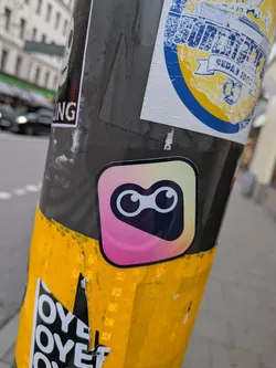 Street sticker The sticker is centrally located and features a rounded square shape with a gradient background transitioning from pink to yellow and then to a light blue. Within this shape, there are two large, circular eyes with black pupils and white irises, giving it a cartoonish and friendly appearance. The sticker has a subtle border.