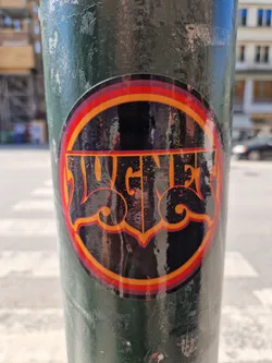 Street sticker A round sticker featuring the word "Jugger" in a stylized, retro font with orange and red accents. The sticker is affixed to a dark green pole and appears glossy.