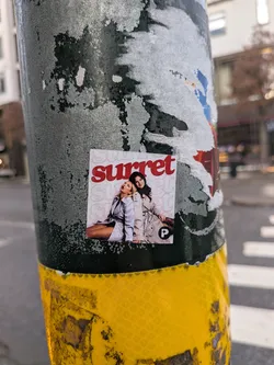 Street sticker A square-shaped sticker featuring two women in stylish trench coats against a light pink background with a faded, repeating 'surret' pattern. The woman on the left has blonde hair, wearing a light grey trench coat, and is captured in a dynamic, open-mouthed pose, looking upwards. The woman on the right has dark hair, wearing a beige trench coat, and is smiling towards the viewer. Above them, the word 'surret' is prominently displayed in bold red, sans-serif lettering. In the bottom right corner, there's a small black square logo with a white circle containing a white 'P'.