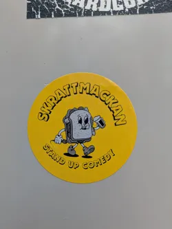 Street sticker A round, yellow sticker featuring a sandwich cartoon character holding a microphone. The text around the character reads "SKRATTMACKAN STAND UP COMEDY".