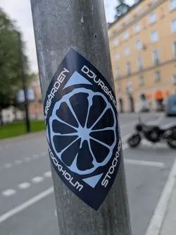 Street sticker The sticker is diamond-shaped and features a stylized, dark blue graphic of a citrus fruit slice with light blue segments. The text "DJURGÅRDEN" is written in white capital letters along the top curve, and "STOCKHOLM" is written along the bottom curve. The background of the sticker appears to be a dark blue, while the fruit and text are in a lighter blue. The sticker is affixed to a grey, cylindrical pole.
