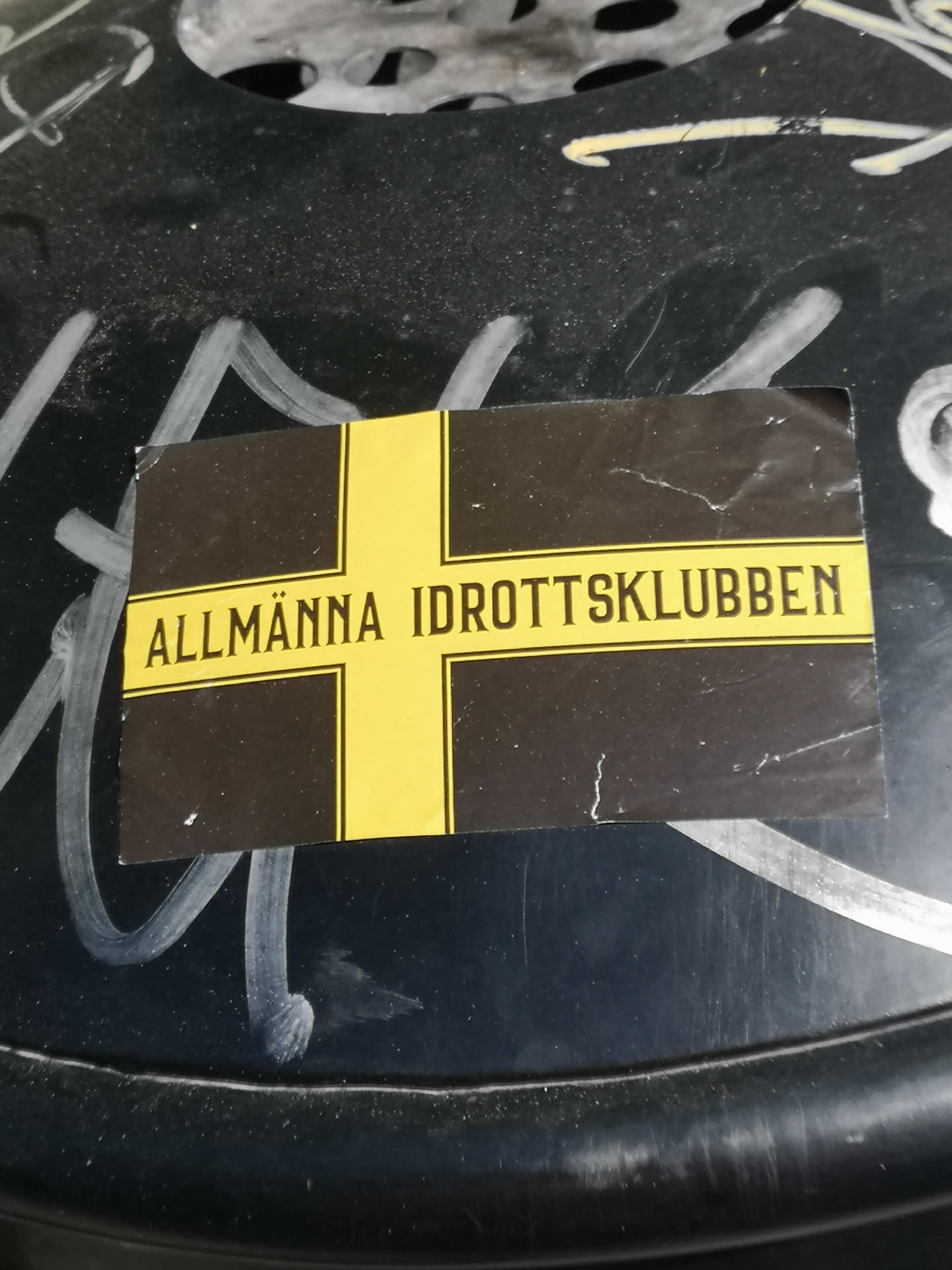 A rectangular sticker featuring a prominent yellow Nordic cross on a black background. The horizontal bar of the cross is wider and contains the text 'ALLMÄNNA IDROTTSKLUBBEN' in black, capitalized, sans-serif letters. The sticker shows signs of wear, including slightly curled corners and minor surface imperfections, suggesting it is a physical sticker adhered to a textured, dark surface with white graffiti.
