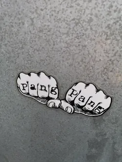 Street sticker A black and white sticker featuring two clenched fists, side-by-side, with knuckles facing forward. Each fist has the word 'Pang' written across its knuckles in a bold, serif-style font. The fists are rendered in a graphic, illustrative style with thick black outlines and cross-hatching for shading, reminiscent of traditional tattoo art or comic book imagery. The sticker has a clear, die-cut border around the white design, making it stand out against the textured, speckled grey background, which appears to be galvanized metal.