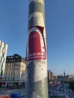 This image shows a sticker affixed to a metal pole. The sticker is mostly maroon with a graphic of a door and the words "Sognuiggio Liberato Laboratorio di Mutuo Soccorso" which translates from Italian to "Liberated Subject Mutual Aid Laboratory."