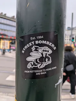 The sticker is black with white text and graphics. At the top, it reads 'Est. 1994' in a curved line. Below that, 'STREET BOMBERS' is written in larger, arched text. The central graphic is a cartoon bee wearing a rugby helmet and holding a rugby ball with 'DRS' written on it. The bee has a determined expression. Below the bee, the text 'FROM ROTTERDAM TO HONG KONG AND BACK' is displayed in two lines. The sticker appears to be slightly worn and peeling at the edges.