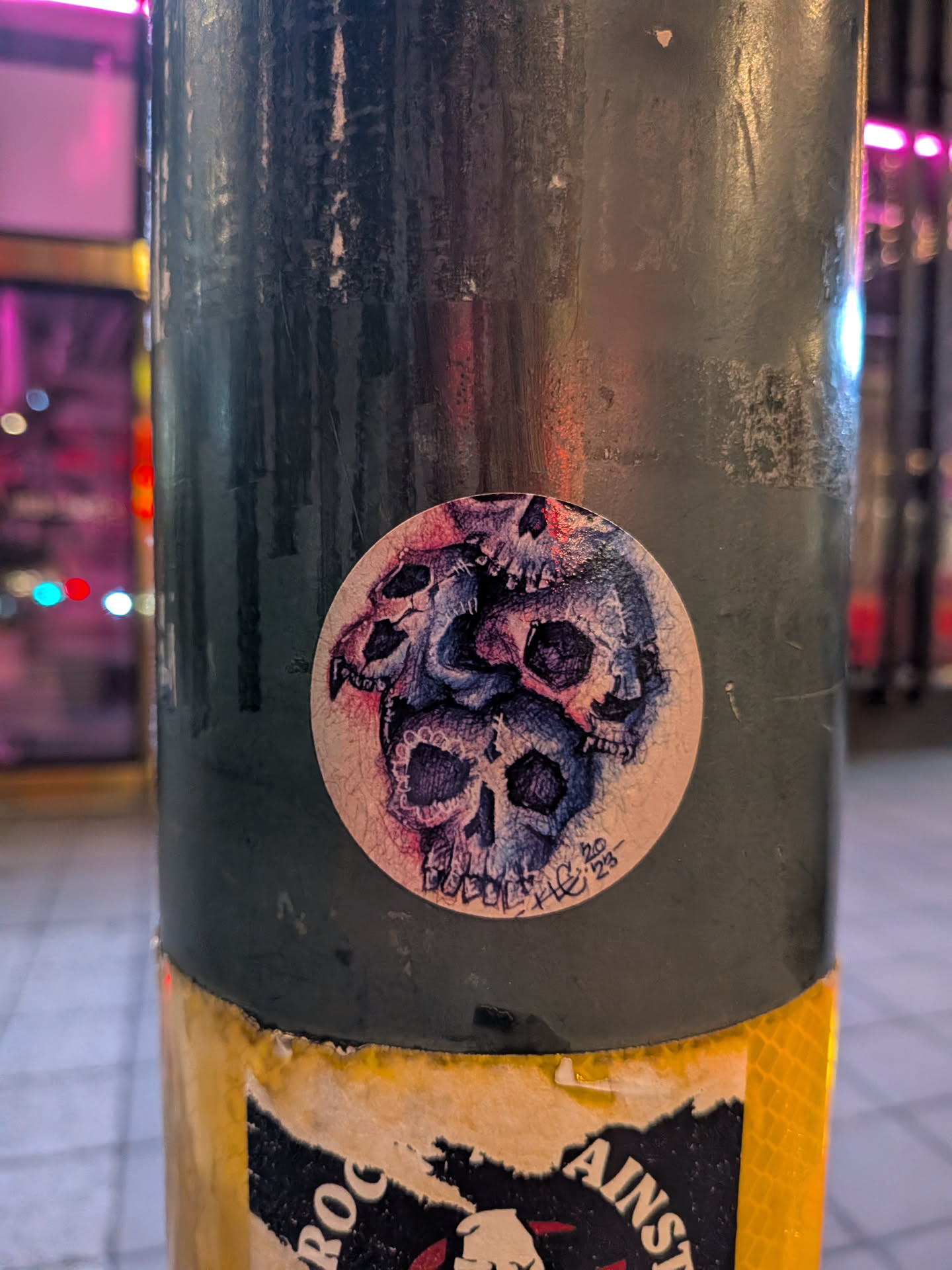 The sticker is circular and features a detailed illustration of multiple skulls overlapping each other. The skulls are depicted in shades of blue and purple, with black outlines and shading to create depth. Some skulls have intricate patterns within their eye sockets and teeth. The overall style appears to be hand-drawn or sketched, with visible lines and texture. In the lower right portion of the sticker, there is some faint text and numbers, possibly a signature and date. The sticker has a slightly worn or textured appearance, suggesting it has been applied for some time.