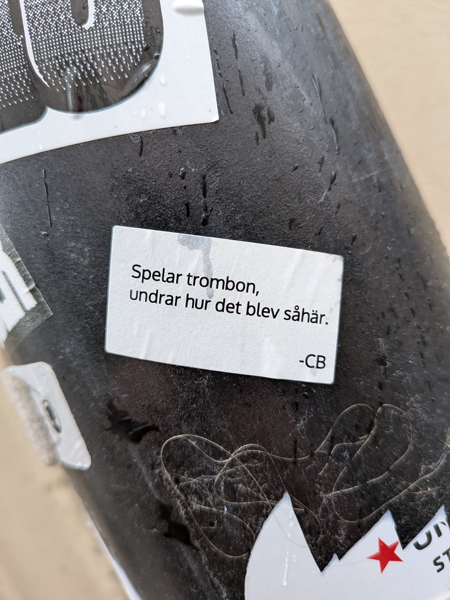 A rectangular white sticker with slightly rounded corners, featuring black sans-serif text. The sticker is applied to a dark, textured surface, possibly metal or plastic, and has several water droplets on its surface, indicating exposure to moisture. The text is centered on the sticker.