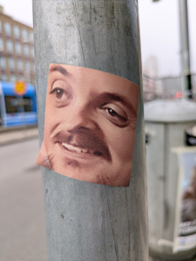 A square sticker affixed to a weathered, gray metal pole displays a close-up image of a man's face. The man has fair skin, brown eyes, and a mustache. His expression appears playful, with his eyes slightly upturned and a faint smile. The background shows an urban environment with buildings and a blurred tram.