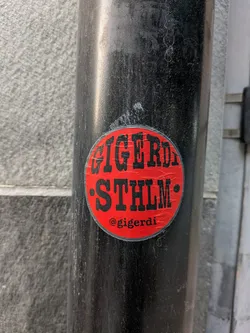 Street sticker The sticker is circular with a bright red background. Bold, black, sans-serif lettering is prominent, spelling out "GIGERD STHLM" across the top two-thirds of the circle. Below this text, centered, is the text "@gigerdi" in a smaller, white font. The sticker appears to be slightly worn, with a few minor scuffs and imperfections on its surface.