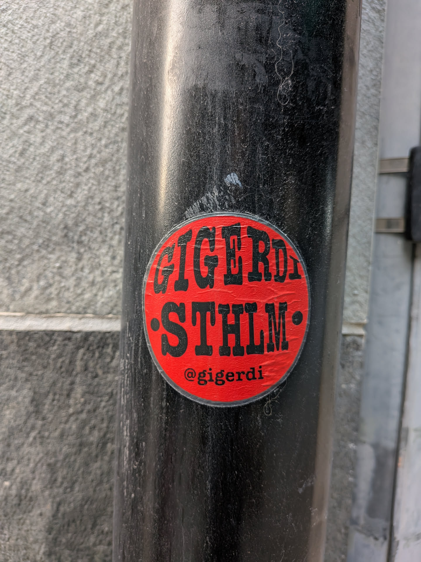 The sticker is circular with a bright red background. Bold, black, sans-serif lettering is prominent, spelling out "GIGERD STHLM" across the top two-thirds of the circle. Below this text, centered, is the text "@gigerdi" in a smaller, white font. The sticker appears to be slightly worn, with a few minor scuffs and imperfections on its surface.