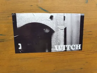 Street sticker A black and white photo sticker featuring an archway made of bricks. The word "WITCH" is printed in white sans-serif font at the bottom right. There's a mysterious, almost eye-like shape visible in the dark archway. The sticker has a slightly worn appearance, suggesting it has been placed outdoors.