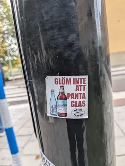 Street sticker A square sticker with a white background, affixed to a black metal pole. It features bold red-brown Swedish text that reads 'GLÖM INTE ATT PANTA GLAS'. To the left of the text are illustrations of two beer bottles: a light blue Tuborg bottle and a brown Eriksberg Pale Ale bottle. In the bottom right corner, there is a circular logo containing the text 'RETUR PANT FLASKA'.