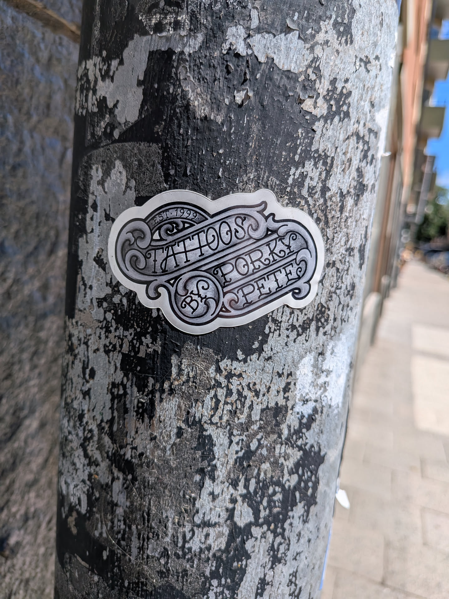 A die-cut sticker featuring ornate, Chicano-style black and grey filigree. The design mimics a classic metallic tattoo shop sign with shaded scrollwork and stylized lettering.