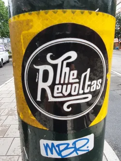 Street sticker A black and yellow sticker featuring the logo for a band called "The Revoltas". There's also a smaller blue sticker underneath it that says MBR.