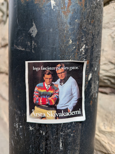 Street sticker This is a sticker featuring two men. The man on the left is wearing a colorful sweater with a tie, and the man on the right is wearing a light blue sweater. The text above them reads "Inga fascister på våra gator", which translates to "No fascists in our streets". Below the image, it says "Årsta Skivakademi".
