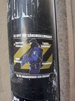 Street sticker The sticker features a central image of a person dressed in a dark blue police uniform, complete with a balaclava covering their face and holding a rifle. This figure is set against a background of yellow and black diagonal hazard stripes. Several white lines with accompanying white text labels point to different aspects of the figure. These labels ironically describe the police officer as 'Aggressiv & Våldsam' (Aggressive & Violent), 'Tungt Beväpnad' (Heavily Armed), 'Typiska Gängkläder' (Typical Gang Clothes), and 'Gängsymbol' (Gang Symbol), with the latter pointing directly to the 'POLIS' (Police) patch on the uniform. A prominent white banner at the top reads 'SE UPP FÖR GÄNGMEDLEMMAR!!!' (Look out for gang members!!!). Below the main image, smaller text states 'BOKTIPS: BATONGERNA SLÅR NEDÅT' (BOOK TIP: THE BATONS STRIKE DOWNWARDS), followed by larger text in quotation marks: '"DE ÄR ORGANISERADE OCH FARLIGA!"' ("THEY ARE ORGANIZED AND DANGEROUS!"). The sticker itself appears to be a physical print, showing some wear and tear, affixed to a dark, textured pole.