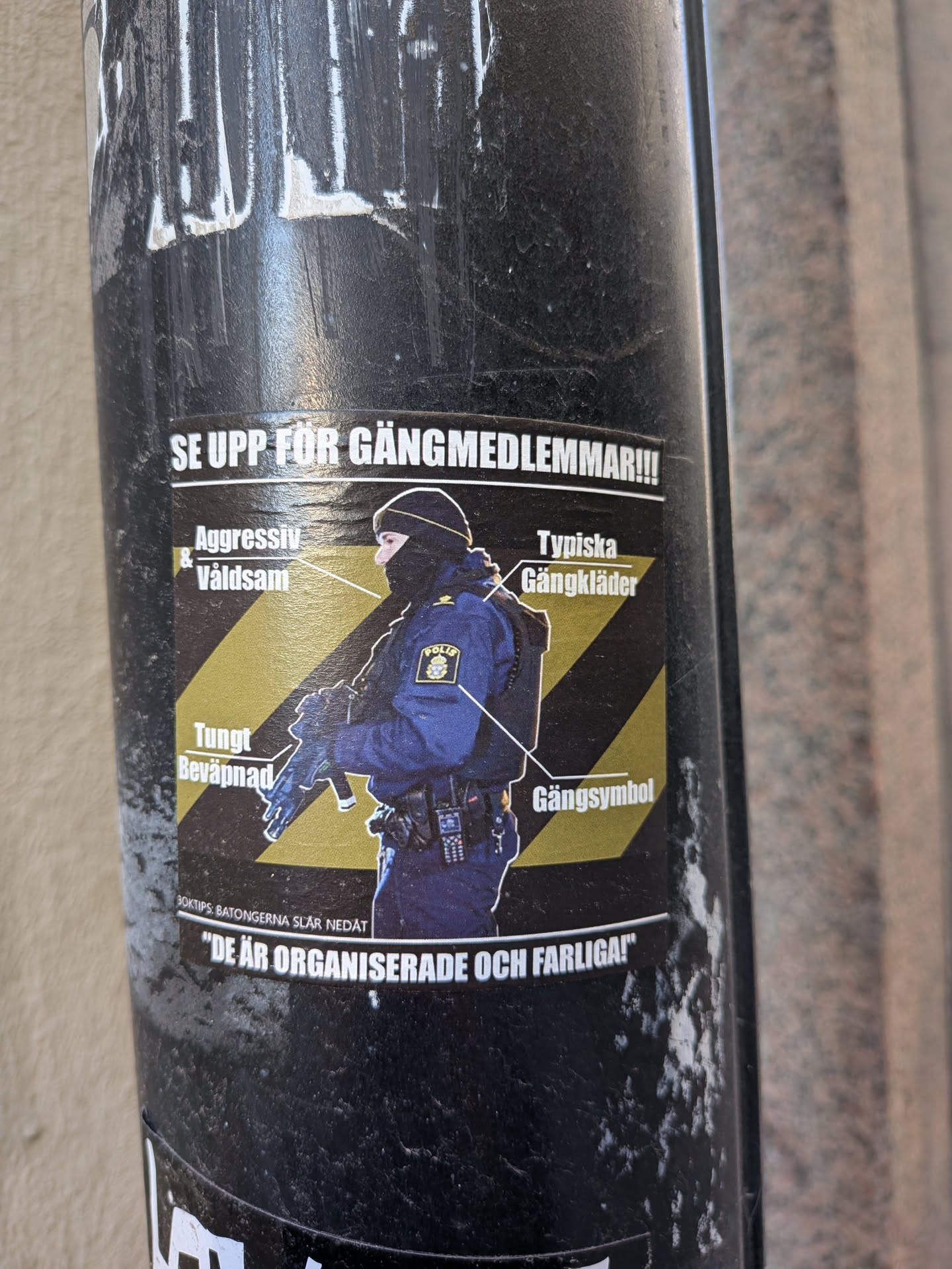The sticker features a central image of a person dressed in a dark blue police uniform, complete with a balaclava covering their face and holding a rifle. This figure is set against a background of yellow and black diagonal hazard stripes. Several white lines with accompanying white text labels point to different aspects of the figure. These labels ironically describe the police officer as 'Aggressiv & Våldsam' (Aggressive & Violent), 'Tungt Beväpnad' (Heavily Armed), 'Typiska Gängkläder' (Typical Gang Clothes), and 'Gängsymbol' (Gang Symbol), with the latter pointing directly to the 'POLIS' (Police) patch on the uniform. A prominent white banner at the top reads 'SE UPP FÖR GÄNGMEDLEMMAR!!!' (Look out for gang members!!!). Below the main image, smaller text states 'BOKTIPS: BATONGERNA SLÅR NEDÅT' (BOOK TIP: THE BATONS STRIKE DOWNWARDS), followed by larger text in quotation marks: '"DE ÄR ORGANISERADE OCH FARLIGA!"' ("THEY ARE ORGANIZED AND DANGEROUS!"). The sticker itself appears to be a physical print, showing some wear and tear, affixed to a dark, textured pole.