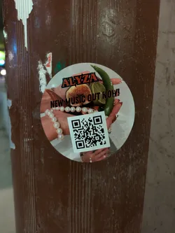 Street sticker This round sticker is promoting new music from Alyza. It features a pair of hands holding fruit and a green pepper, adorned with rings and a pearl bracelet.  The sticker also includes a QR code.