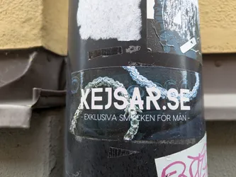 Street sticker The sticker is predominantly dark with white lettering. Prominently displayed in large white font is 'KEJSAR.SE'. Below this, in smaller white text, reads '- EXKLUSIVA SMYCKEN FÖR MÄN -'. The lettering is overlaid on a dark background that features a detailed image of a thick, intertwined silver-colored chain with blueish highlights, suggesting luxury and craftsmanship. The sticker is slightly curved, conforming to the cylindrical surface it's adhered to, and shows some signs of wear, with minor peeling and scuffing at the edges and surface.