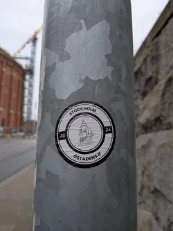 The sticker is circular with a black and white design. It features a detailed illustration of a sailing ship in the center, with waves beneath it. The outer ring of the sticker contains text. At the top, it reads 'STOCKHOLM', and at the bottom, it reads 'ÖSTADENS IF'. The numbers '20' and '25' are positioned on the left and right sides of the outer ring, respectively. The sticker has a slightly worn appearance, with visible creases and texture, suggesting it is indeed a physical sticker applied to the pole.