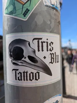 This is a sticker for Tris Blu Tattoo. It features a grayscale illustration of a bird skull on the left, and the text "Tris * Blu Tattoo" to the right of and below the skull in a gothic-style font. The background is white with a thin green border and a dotted blue line running inside the green border.