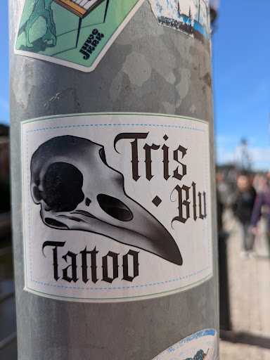 This is a sticker for Tris Blu Tattoo. It features a grayscale illustration of a bird skull on the left, and the text "Tris * Blu Tattoo" to the right of and below the skull in a gothic-style font. The background is white with a thin green border and a dotted blue line running inside the green border.