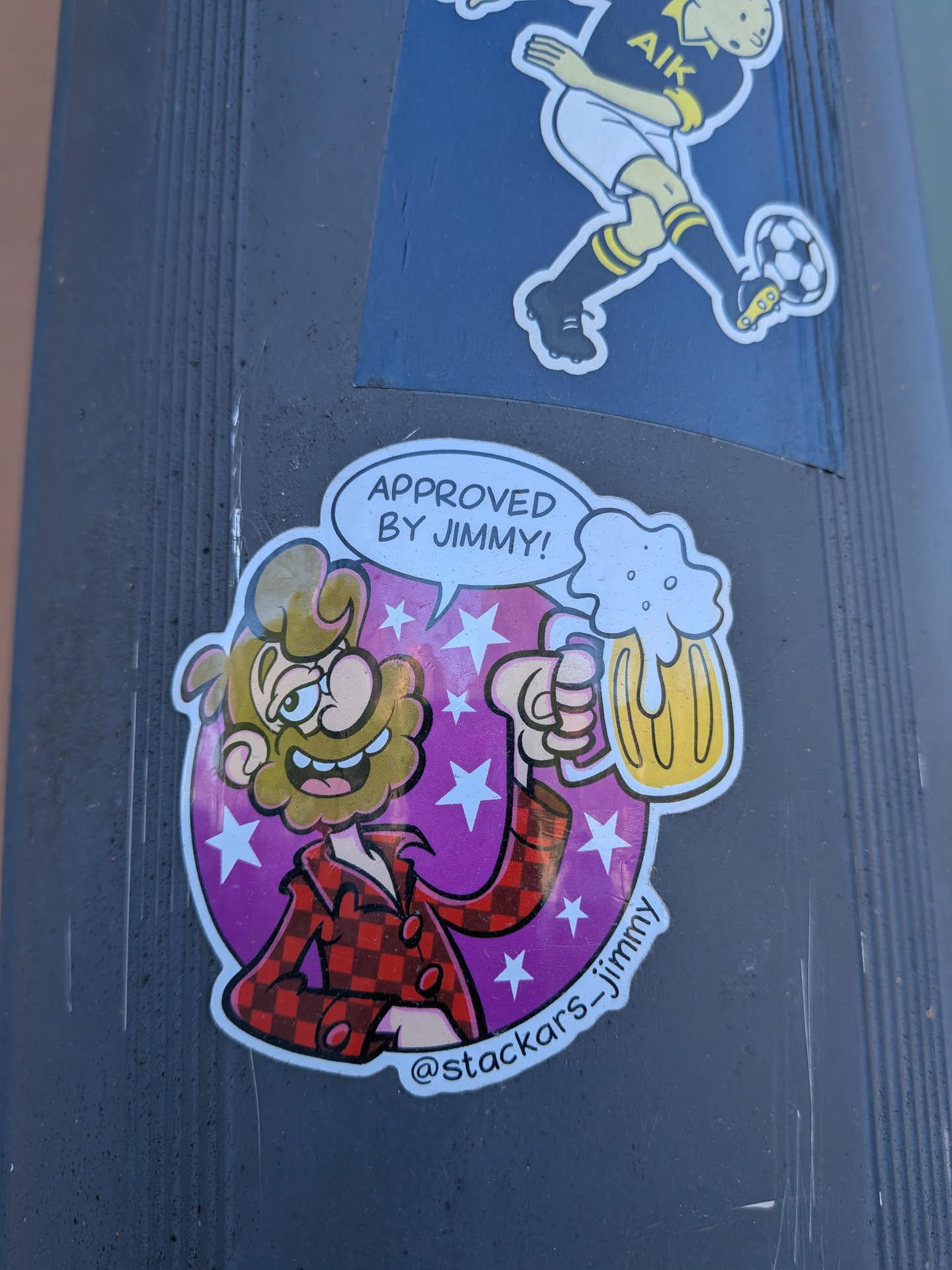 A vibrant, die-cut sticker featuring a cartoon man with a large, bushy brown beard and hair, a wide smile, and big eyes. He is wearing a red and black plaid shirt and holding up a large, frothy mug of golden beer in his right hand. Above his head, a white speech bubble contains text. The character is set against a bright purple background adorned with numerous white stars of varying sizes. The sticker has a playful, somewhat rustic yet modern cartoon aesthetic, with a social media handle visible at the bottom.