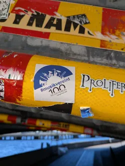 Street sticker A square sticker with a white background and a blue semicircular design at the top. The blue area contains a silhouette of various musical instruments, including a trumpet, saxophone, clarinet, and a conductor's baton. Below the instruments, the text '#musikverein' is prominently displayed in blue, followed by the number '100' in a larger, bold font. Beneath the '100', the text 'MUSIKVEREIN' and 'Langenau 1925' are written in smaller black font. The sticker appears to be slightly creased and adhered to a yellow, textured surface.