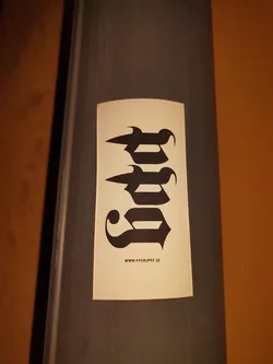 A beige sticker with black stylized lettering, possibly the name "JUD",  and a website address at the bottom. The style is reminiscent of gothic lettering or a custom typeface. The sticker is affixed to a dark grey cylindrical surface.