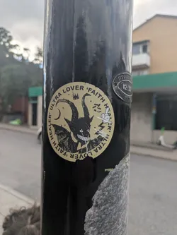 Street sticker The sticker is circular, featuring a bold black illustration on a muted gold or tan background. The central image is a stylized, horned creature with large, curved horns, bat-like wings, and a wide, toothy grin revealing sharp, white teeth. Its eyes are small, glowing white dots. Above its head, a small crown is visible. To the left, a crescent moon and several small stars are depicted. To the right, a cartoonish white hand with a pointing finger emerges, surrounded by jagged lines resembling lightning bolts. The text "ULTRA LOVER 'FAITH HEALER'" is repeated twice, forming a circular border around the central illustration. The sticker shows some wear, particularly at its bottom edge.