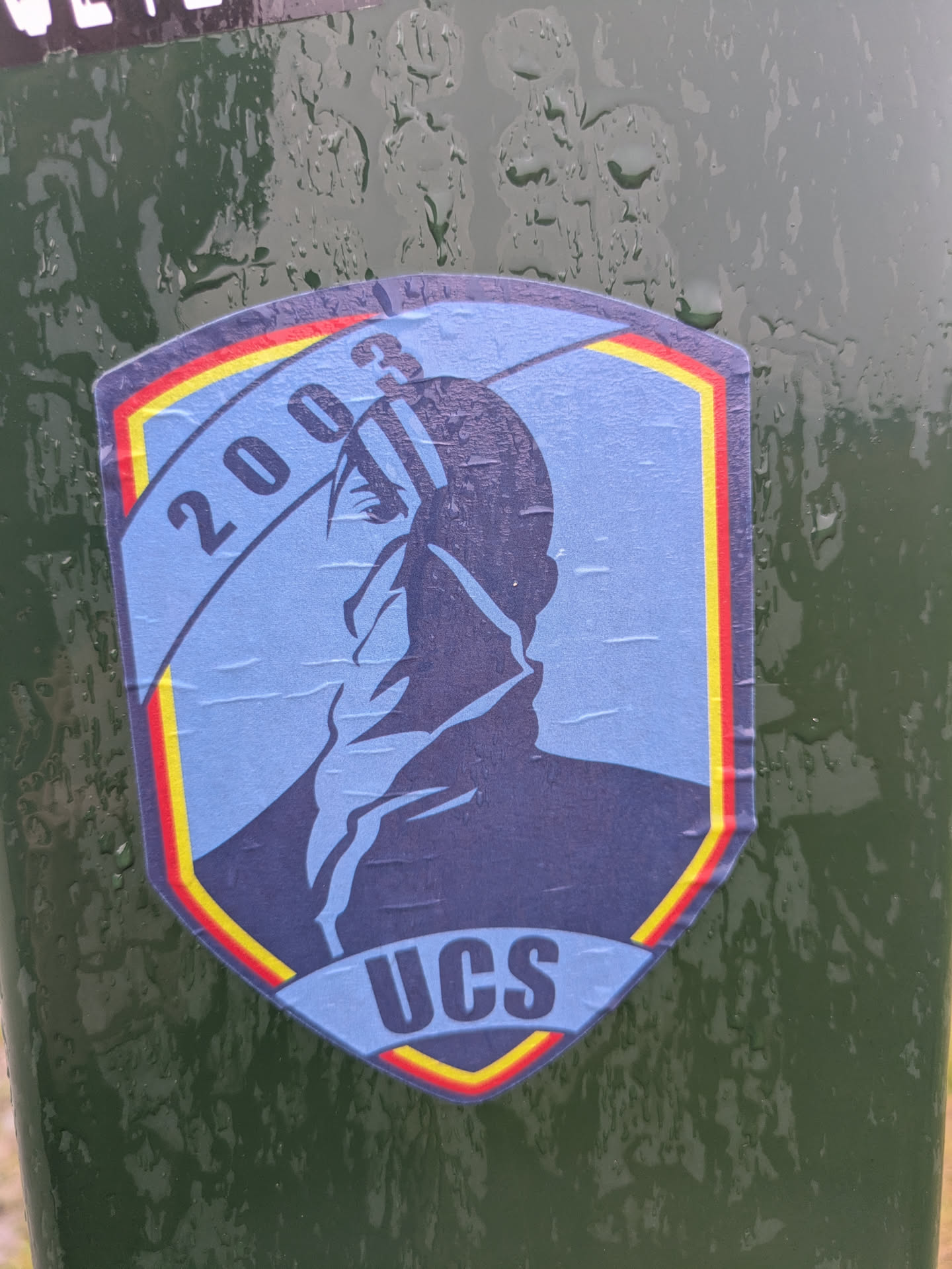 Street sticker A shield-shaped sticker adhered to a wet, dark green surface. The sticker has a light blue background with a dark blue silhouette of a person's head and shoulders in profile, facing left. The person appears to be wearing a high-collared garment. A curved banner crosses the upper left, displaying the numbers '2003'. At the bottom center, the letters 'UCS' are prominently displayed. The sticker is bordered by concentric lines of red, yellow, and a darker blue. The surface of the sticker shows visible creases and is covered in water droplets, indicating it is a physical sticker exposed to the elements.
