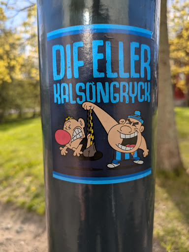 The sticker, attached to a pole, features a cartoonish scene with the text "DIF ELLER KALSONGRYCK". It depicts one character pulling up the underwear of another character, causing the victim to grimace in discomfort. The style is reminiscent of classic cartoon humor.