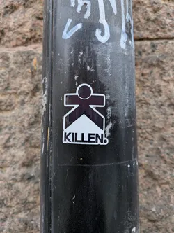 Street sticker A die-cut sticker affixed to a black metal post. The sticker depicts a stylized character whose body is shaped like a bold letter 'K' with a circular head, rendered in dark brown/black. A white triangular wedge points upward into the lower half of the character. Below the icon, the word 'KILLEN!' is printed in bold black capital letters. The sticker is outlined with a clean white border.