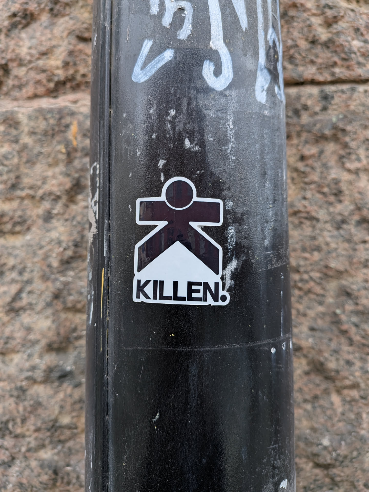 A die-cut sticker affixed to a black metal post. The sticker depicts a stylized character whose body is shaped like a bold letter 'K' with a circular head, rendered in dark brown/black. A white triangular wedge points upward into the lower half of the character. Below the icon, the word 'KILLEN!' is printed in bold black capital letters. The sticker is outlined with a clean white border.